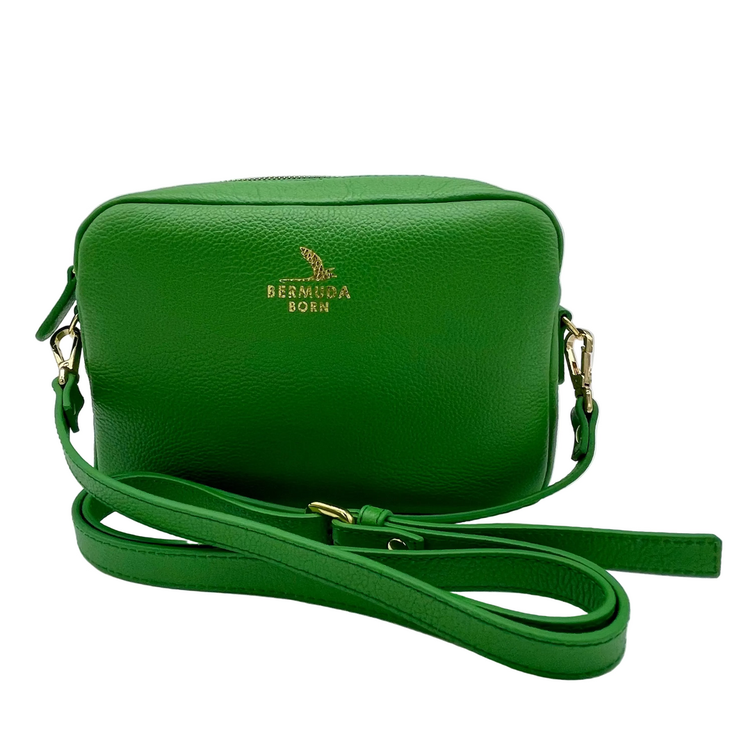 A green textured leather crossbody camera bag with gold hardware and a gold-embossed 'BERMUDA BORN' logo on the front, displayed on a white surface.